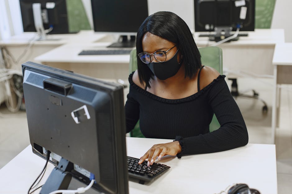 African woman wearing a face mask and eyeglasses working on a computer indoors, ensuring safety and protection.